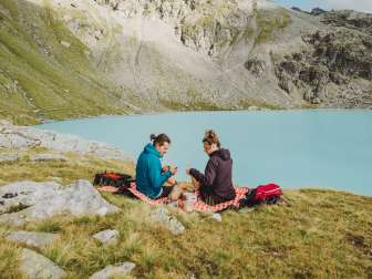 Pärchen sitzt auf einer Picknickdecke vor einem eisblauen Bergsee