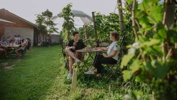 Zwei Personen sitzen inmitten von Weinreben und geniessen gemeinsam ein Glas Wein.
